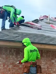 Roofing crew installing shingles beside wrapped material bundles on a residential roof.
